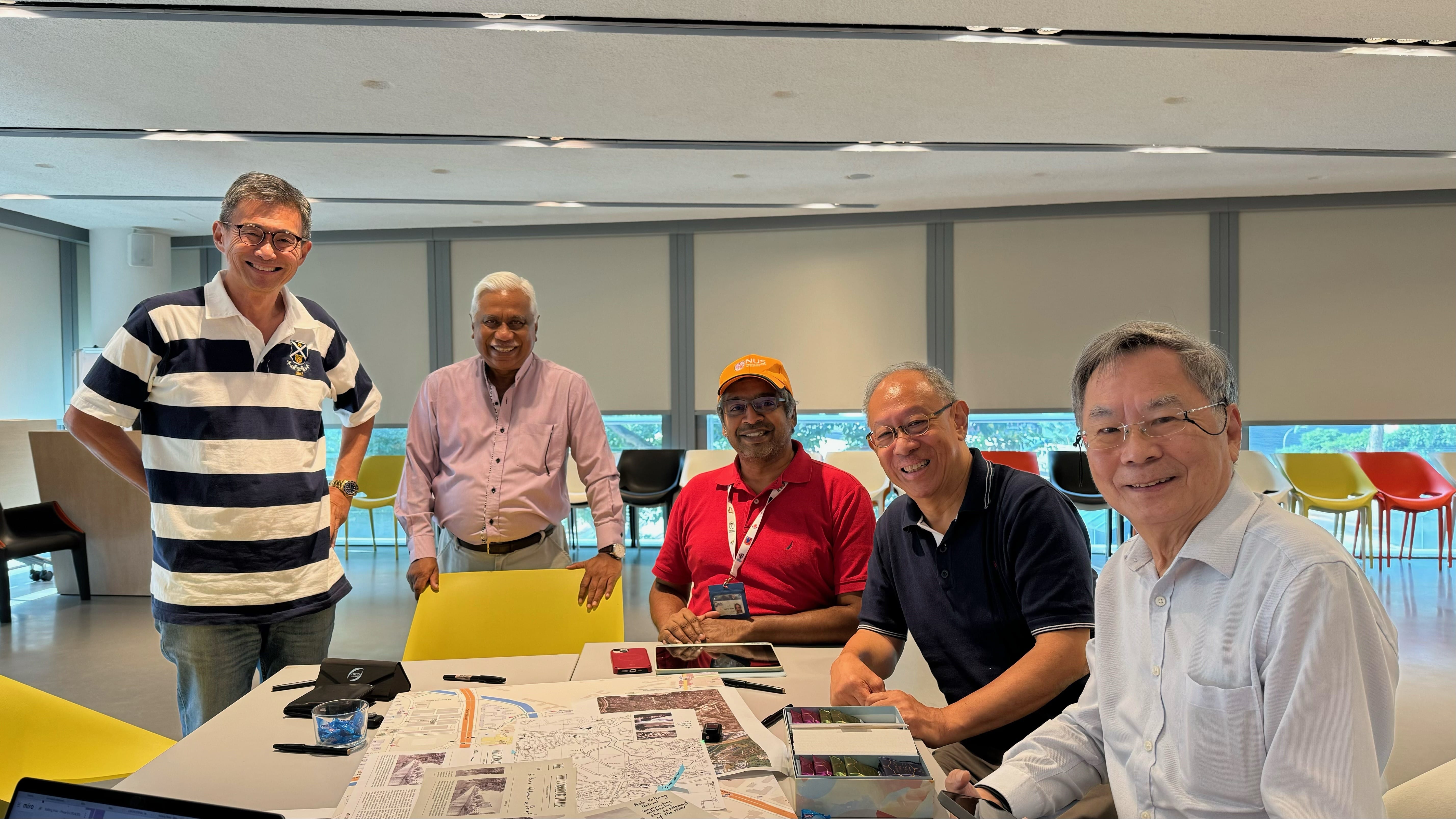 Five smiling men gathered around a table with maps and game pieces in a modern office space.